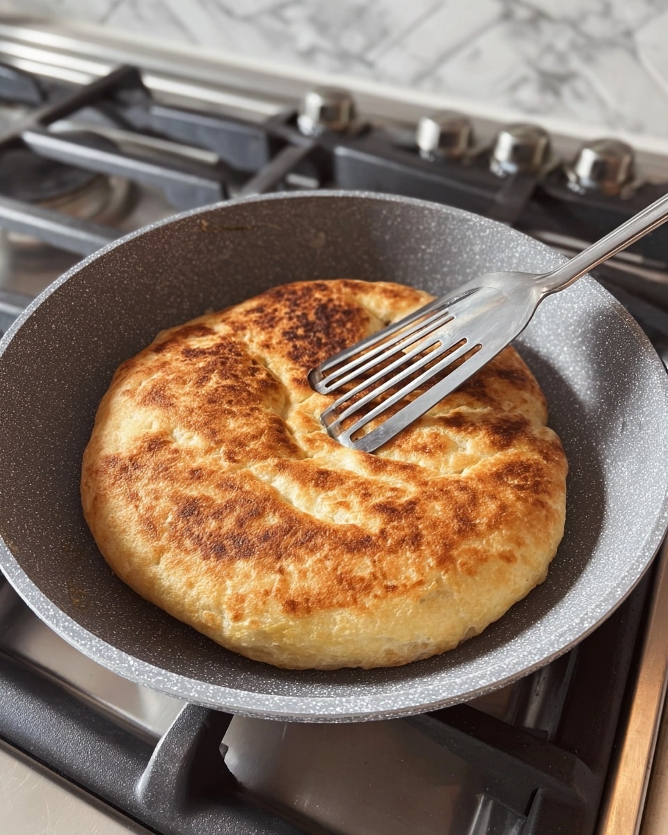 A single round bread is cooking in a gray speckled frying pan on a stove with black grates and silver surface. The bread is golden brown with a slightly uneven texture, showing a crispy crust with some darker brown spots, and a puffy, soft look around the edges. A metal spatula with slits is being used to lift or press the bread gently from the right side of the pan. The background shows the stove knobs and part of the kitchen counter with a white marbled texture. photo taken with an iphone --ar 4:5 --v 7
