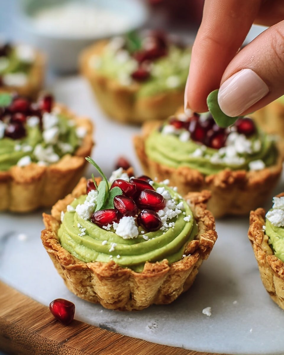 Six small tartlets with a light brown crispy crust are placed on a dark wooden board on a white marbled surface. Each tartlet has three visible layers: the bottom crust, a thick, creamy light green layer swirled on top, and scattered bright red pomegranate seeds with white crumbly cheese sprinkled over both. Fresh green mint leaves are placed on or near some tartlets with a few pomegranate seeds and leaves scattered around. In the background, there is a white bowl filled with pomegranate seeds and a halved lemon. Photo taken with an iphone --ar 4:5 --v 7