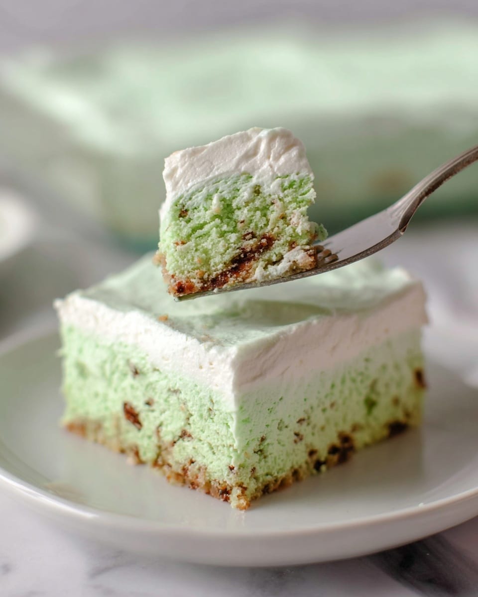 A close-up of a white plate holding a square slice of light green cake with small brown bits inside. The cake has two layers: a moist, crumbly green base with brown specks and a thick, smooth white cream layer on top that looks soft and fluffy. In the foreground, a piece of the cake is lifted on a silver fork, showing the texture of the green base and the creamy white top. The background features a white marbled surface with soft, blurry elements. photo taken with an iphone --ar 4:5 --v 7