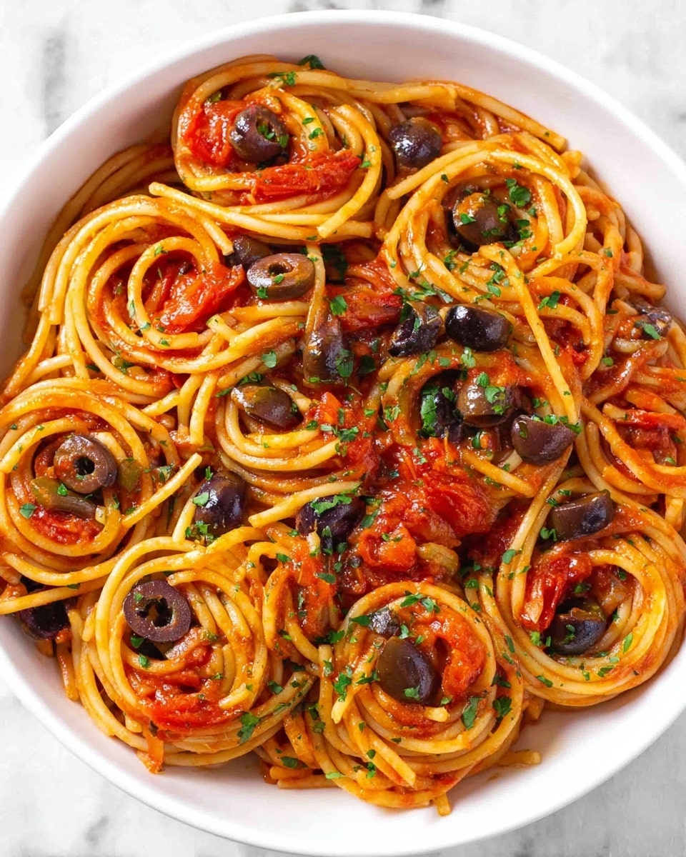 A white bowl filled with spaghetti pasta arranged in small circular nests layered evenly inside. The spaghetti is coated with a shiny, red tomato sauce that has small chunks and bits of dark brown olives and green herbs mixed throughout. Sprinkle of finely chopped green parsley is scattered over the top, adding a fresh touch. The bowl is set on a white marbled surface with a fork placed to the upper right side and a small glass bowl of mixed green and black olives partially visible to the upper left. Photo taken with an iphone --ar 4:5 --v 7