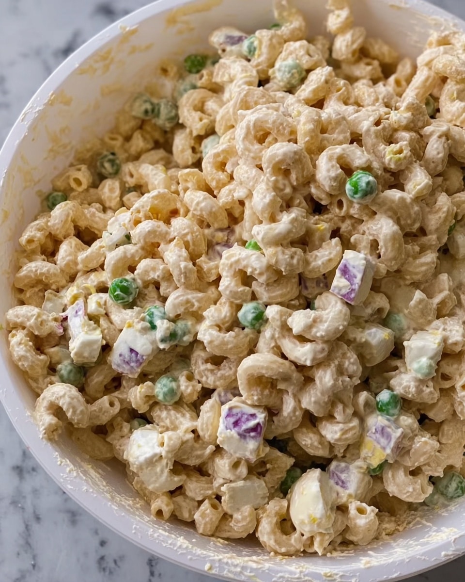 The image shows a close-up of a large white bowl filled with creamy macaroni salad. The salad has small elbow macaroni coated in a thick, pale beige dressing. Mixed in are small pieces of chopped hard-boiled egg, green peas, and bits of onions scattered throughout, adding touches of white, green, and purple to the dish. The texture looks rich and smooth with visible chunks from the added ingredients. The bowl sits on a white marbled surface. photo taken with an iphone --ar 4:5 --v 7