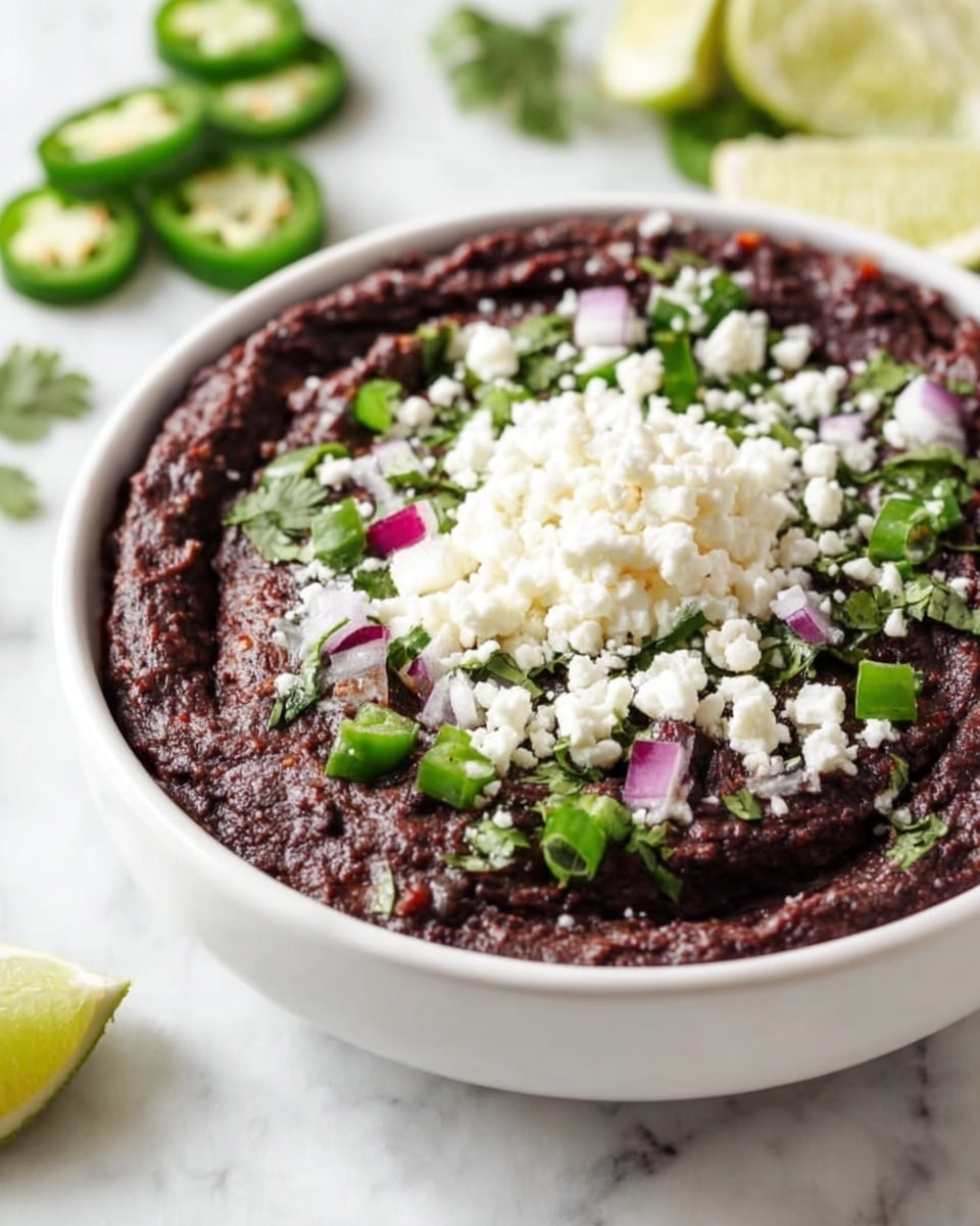 The image shows a white bowl filled with dark purple black bean dip as the bottom layer, which has a smooth but textured surface. On top of this, there are small pieces of green chopped jalapeño, red chopped onion, and fresh green cilantro leaves scattered evenly. The center is topped with a generous mound of white crumbled cheese, creating a contrast against the darker dip. In the background on the white marbled surface, there are sliced green jalapeños and lime wedges slightly out of focus. photo taken with an iphone --ar 4:5 --v 7