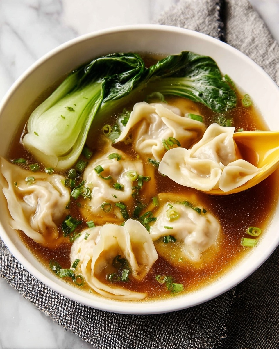 A white bowl filled with clear brown broth, containing six light beige dumplings with soft, folded textures floating in the soup. On one side of the bowl, there are two pieces of bright green bok choy with visible leafy veins, partially submerged. Small green chopped herbs are sprinkled on the dumplings and broth. A yellow spoon is partially dipped into the soup, holding one dumpling. The bowl rests on a grey cloth over a white marbled surface. Photo taken with an iphone --ar 4:5 --v 7