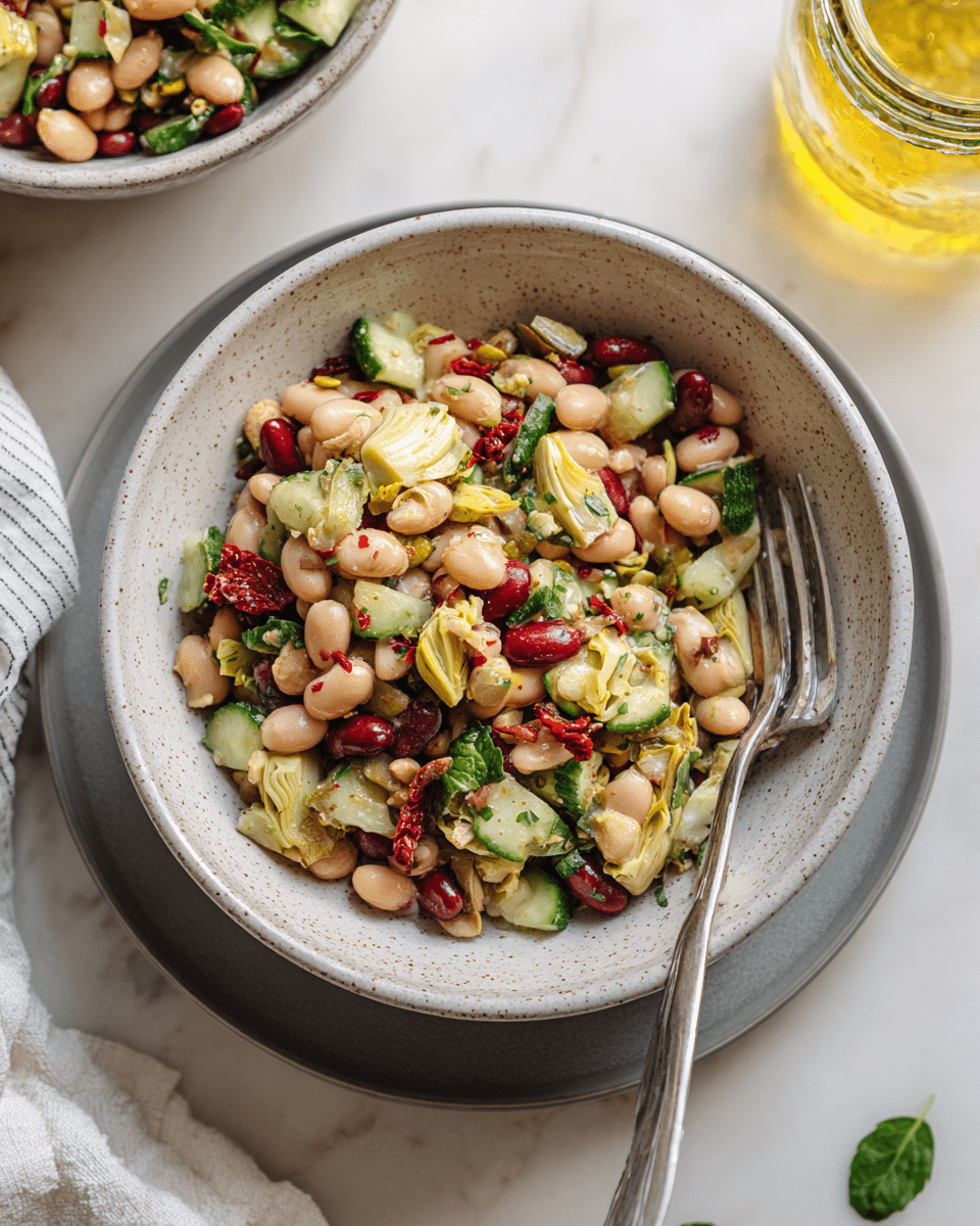 A white speckled bowl filled with a colorful bean salad sits on a white plate, which is placed on a white marbled surface. The salad contains three visible layers: a base layer of beige chickpeas and beige white beans, a middle layer of sliced green cucumber and red kidney beans, and a top layer of chopped fresh green herbs and bits of sun-dried tomato, creating a mix of green, red, beige, and white colors. A silver fork rests inside the bowl, angled towards the viewer. In the background, a partially visible salad on a white plate and a glass jar wrapped in a striped cloth can be seen. Fresh green parsley leaves lie on the marble surface near the bowl. Photo taken with an iphone --ar 4:5 --v 7