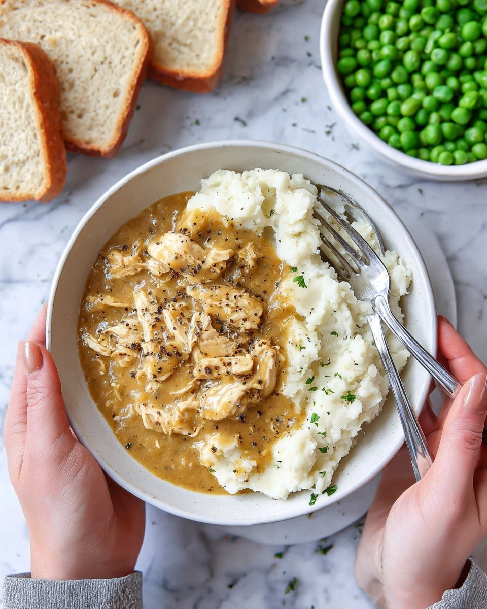 A white bowl held by two woman's hands contains a dish with two main layers: the bottom layer is creamy white mashed potatoes with a soft, smooth texture, and the top layer is shredded chicken covered in a golden brown gravy with visible black pepper specks. Two silver forks rest inside the bowl across the mashed potatoes and chicken. In the upper background, there is a white bowl filled with bright green peas, and to the left, several slices of light brown bread. The surface beneath the bowl is a white marbled texture. photo taken with an iphone --ar 4:5 --v 7