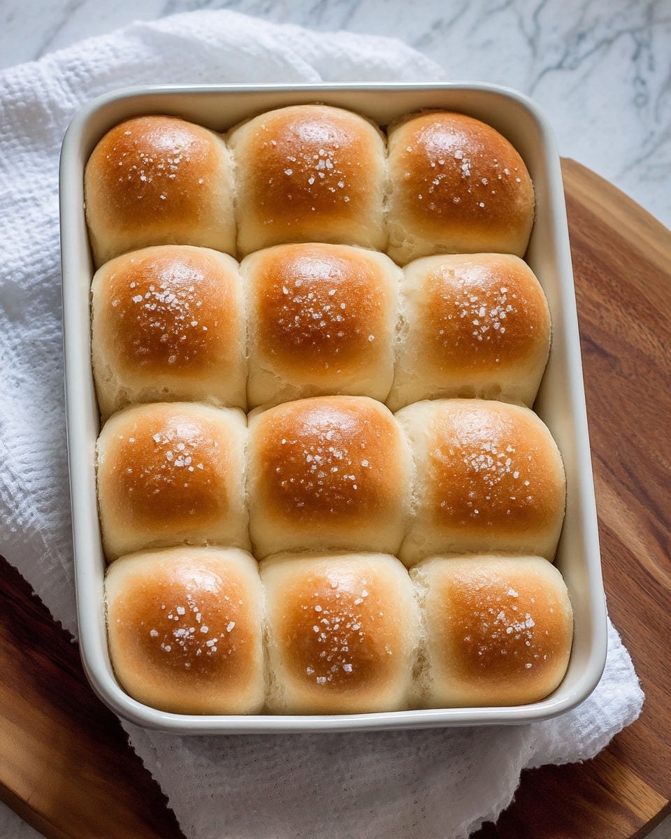 A close-up of a woman's hand holding a soft, light brown bread roll with a fluffy, airy inside texture visible where a piece is torn off. The bread roll is shown in front of a blurred background with more similar bread rolls placed on a white bowl. The overall scene has a warm, soft focus with natural lighting and a white marbled texture behind. photo taken with an iphone --ar 4:5 --v 7