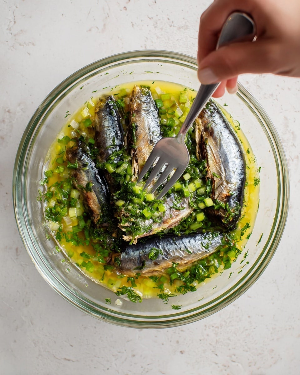 A clear glass bowl sits on a white marbled texture, filled with several whole sardines with shiny, silvery-black skin, arranged in a circle. Around the fish are chopped green herbs and green onion pieces, adding a fresh touch of vivid green. A yellow, oily sauce with small bubbles covers the bottom inside the bowl, blending with the herbs and fish. A woman's hand holds a silver fork pressing down on the sardines in the center, showing a mixing action. photo taken with an iphone --ar 4:5 --v 7