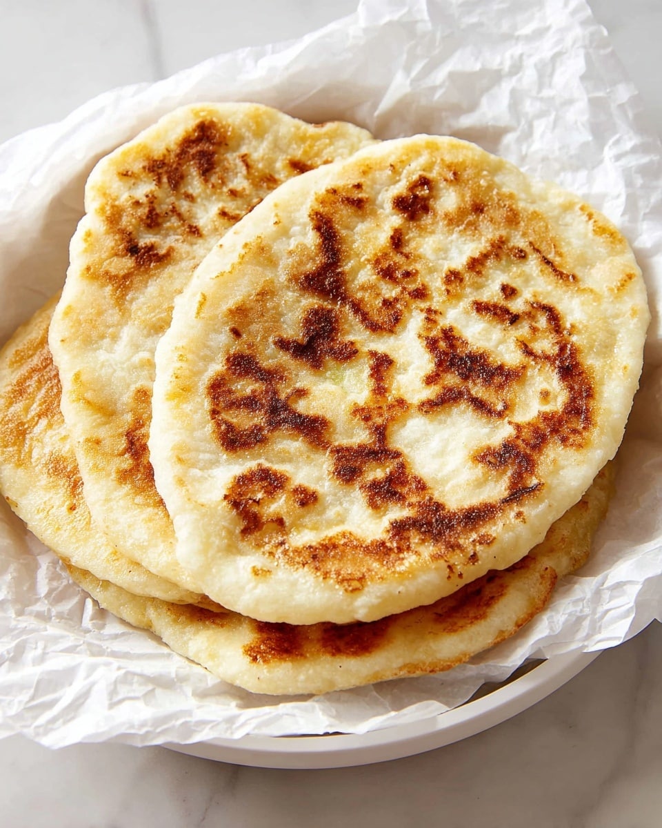 A white round plate holds three thick, golden brown flatbreads stacked on top of each other, with crispy, uneven browned patches on their soft, fluffy surfaces. The flatbreads are placed on crumpled white parchment paper that adds texture to the scene. The background is a white marbled texture that gives a clean, bright feel to the image. The flatbreads are large and slightly irregular in shape, with a light, airy texture visible along the edges. photo taken with an iphone --ar 4:5 --v 7
